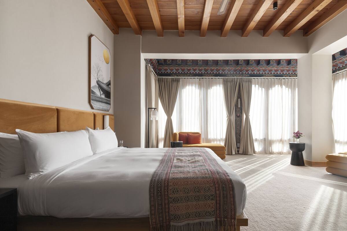 Quarto de hotel com uma cama grande, cabeceira de madeira, um sofá pequeno e uma janela com cortinas estampadas.