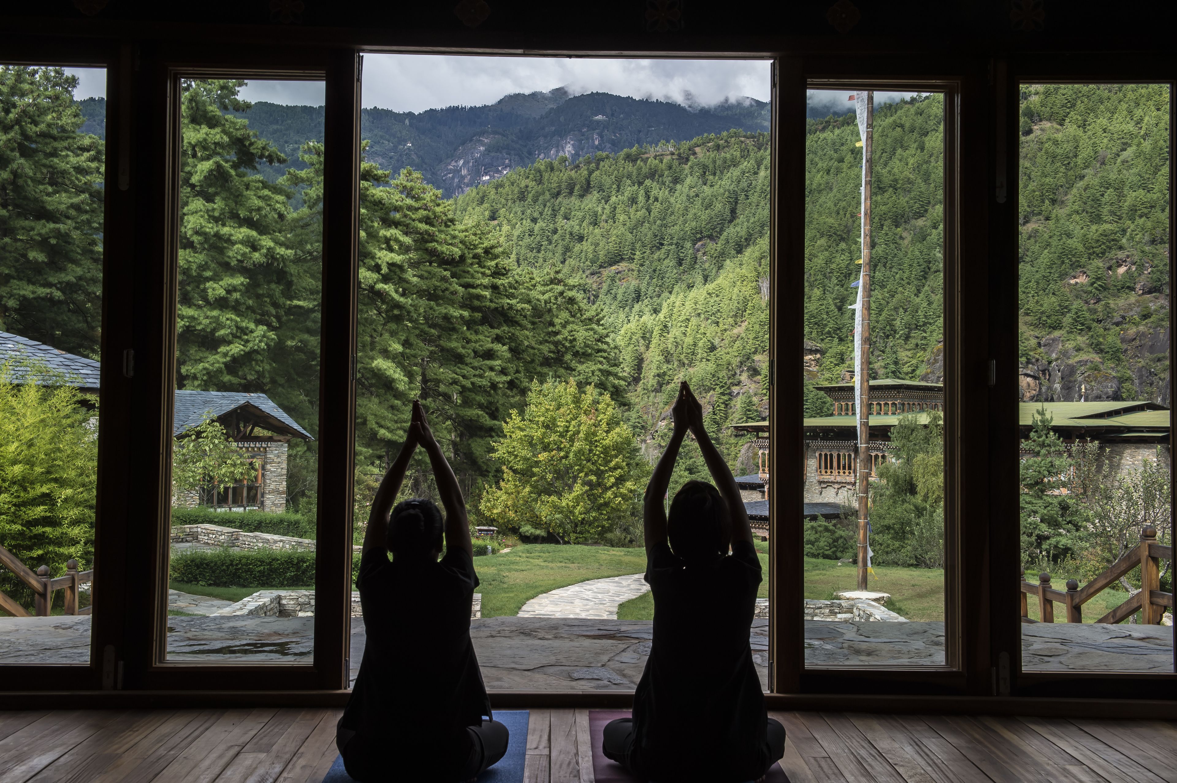 Two people doing yoga in a room with large windows overlooking a mountainous landscape.