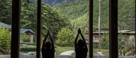 Two people doing yoga in a room with large windows overlooking a mountainous landscape.