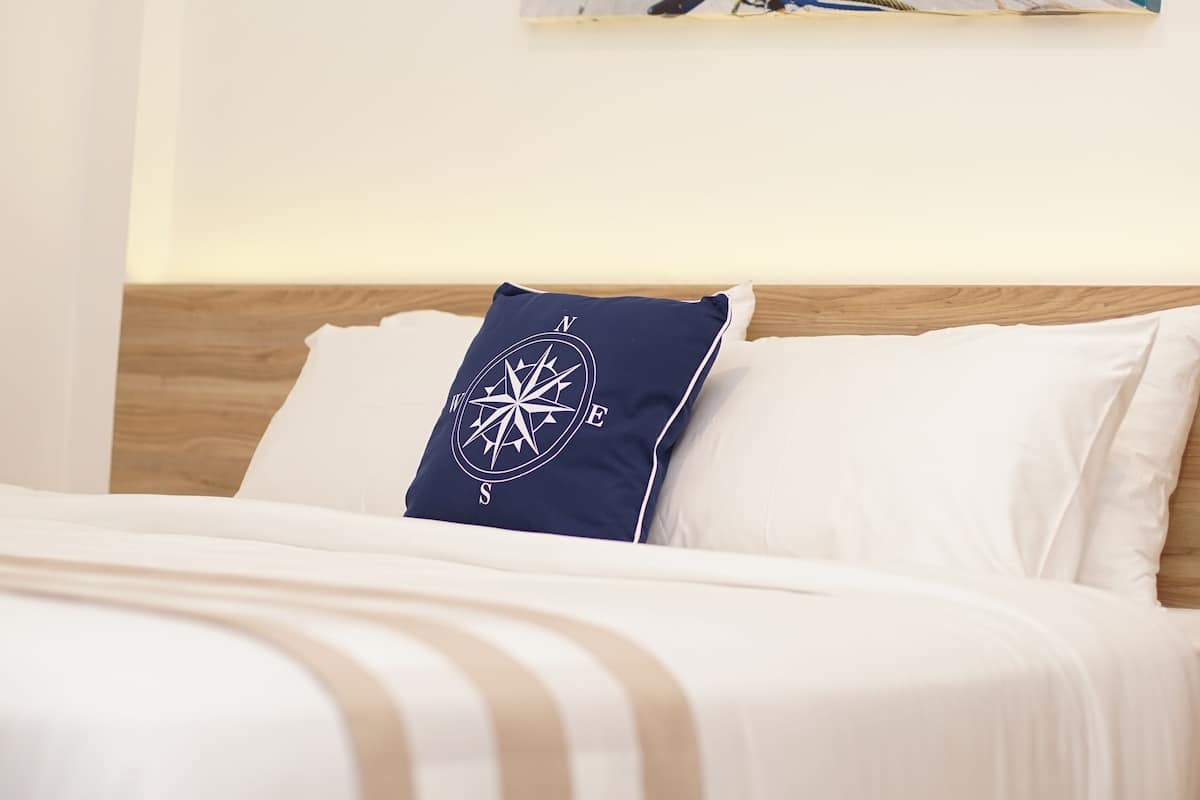 Una cama con ropa de cama a rayas blancas y beige, una almohada con forma de brújula y un cuadro de un barco en un muelle con una isla tropical al fondo.