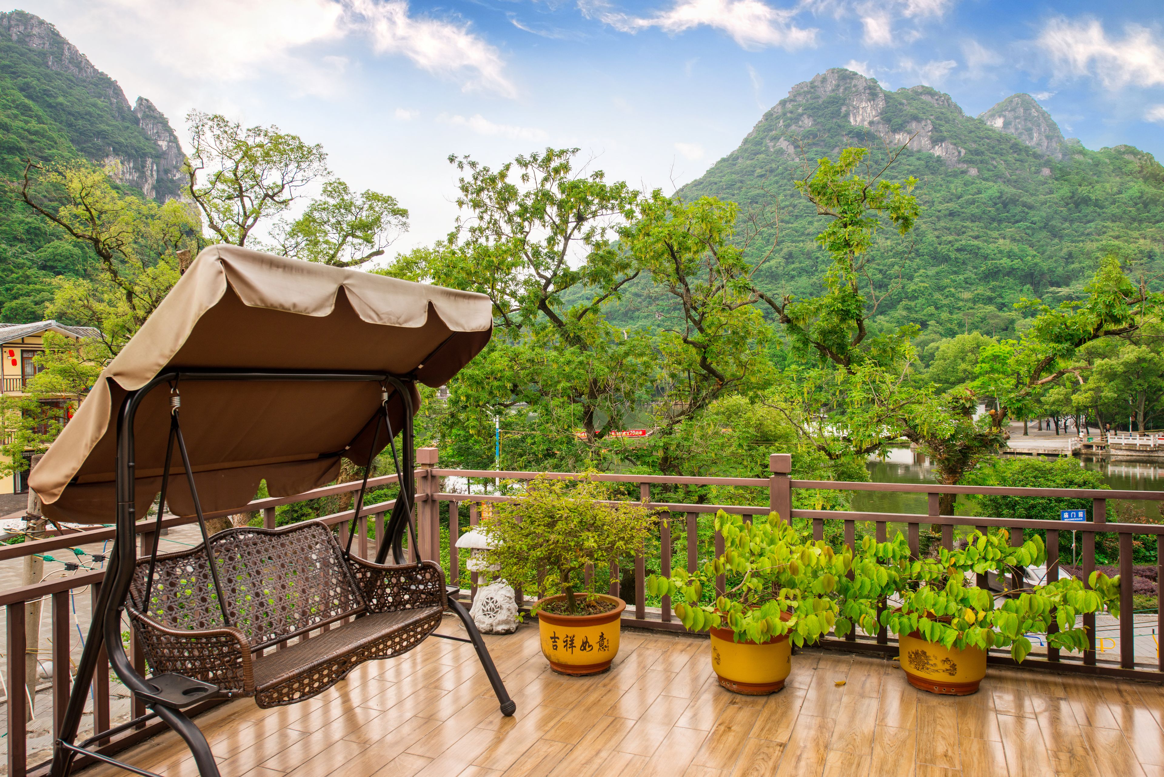A wooden deck with a swing chair, potted plants, and a view of mountains and a lake.