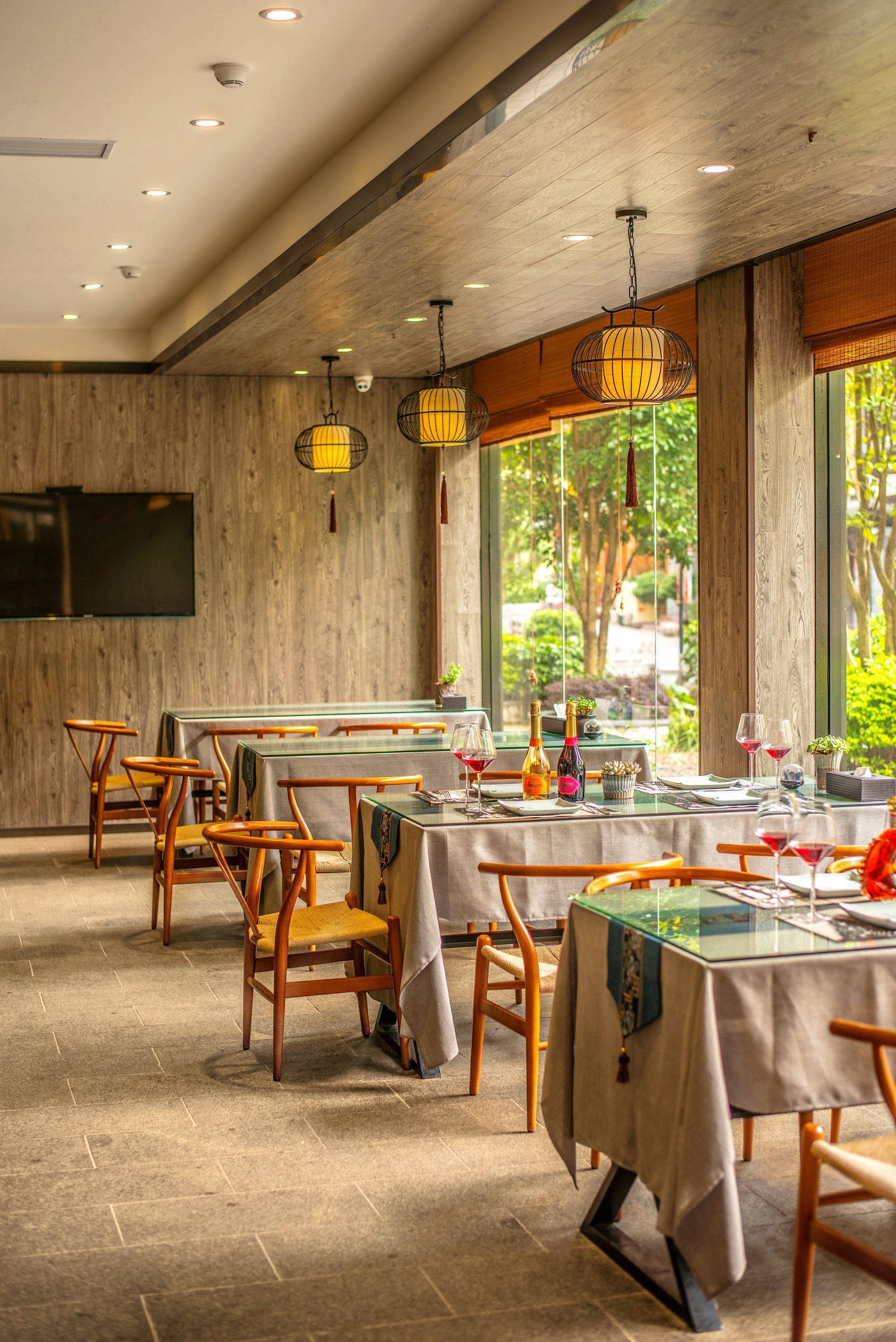 A restaurant with wooden walls, hanging lanterns, and a view of greenery outside. Tables are set with wine glasses and bottles.