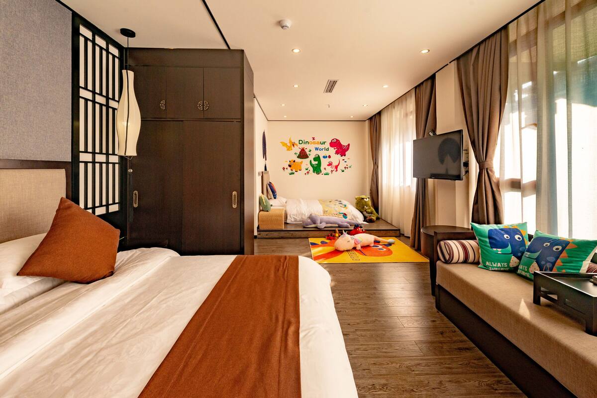 Quarto de hotel com uma cama grande, um sofá, uma TV e uma parede com um mural de 'Dinosaur World'.