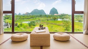 Una habitación con un ventanal grande que da a un paisaje exuberante, con montañas y un río. Hay una mesa trenzada con un libro y un tazón encima, y dos otomanas tejidas.