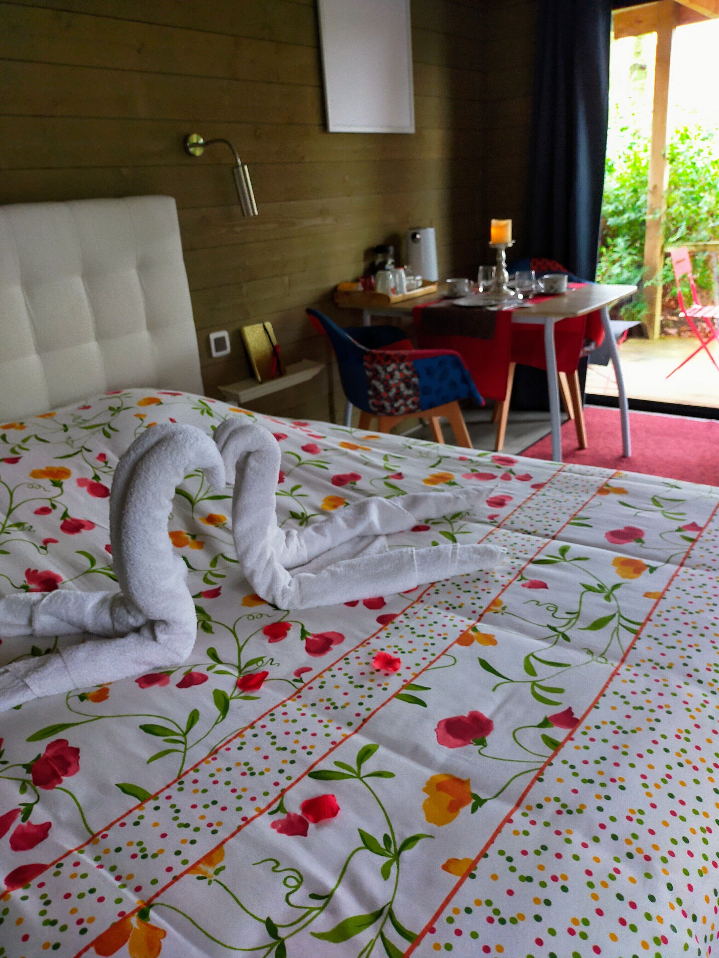 A neatly made bed with a floral bedspread and a towel arranged in a heart shape. There is a wooden headboard, a small bedside table with a lamp, and a window with a view of greenery outside.