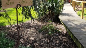 A wooden sign with the text "Les îlots" in a garden area with a wooden deck, surrounded by trees and plants. - Le Clos du Clocher (Gueudecourt)