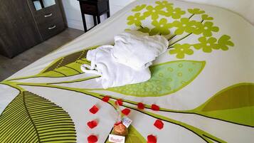 Una cama con una colcha estampada con hojas, una toalla blanca y pétalos de rosa roja.