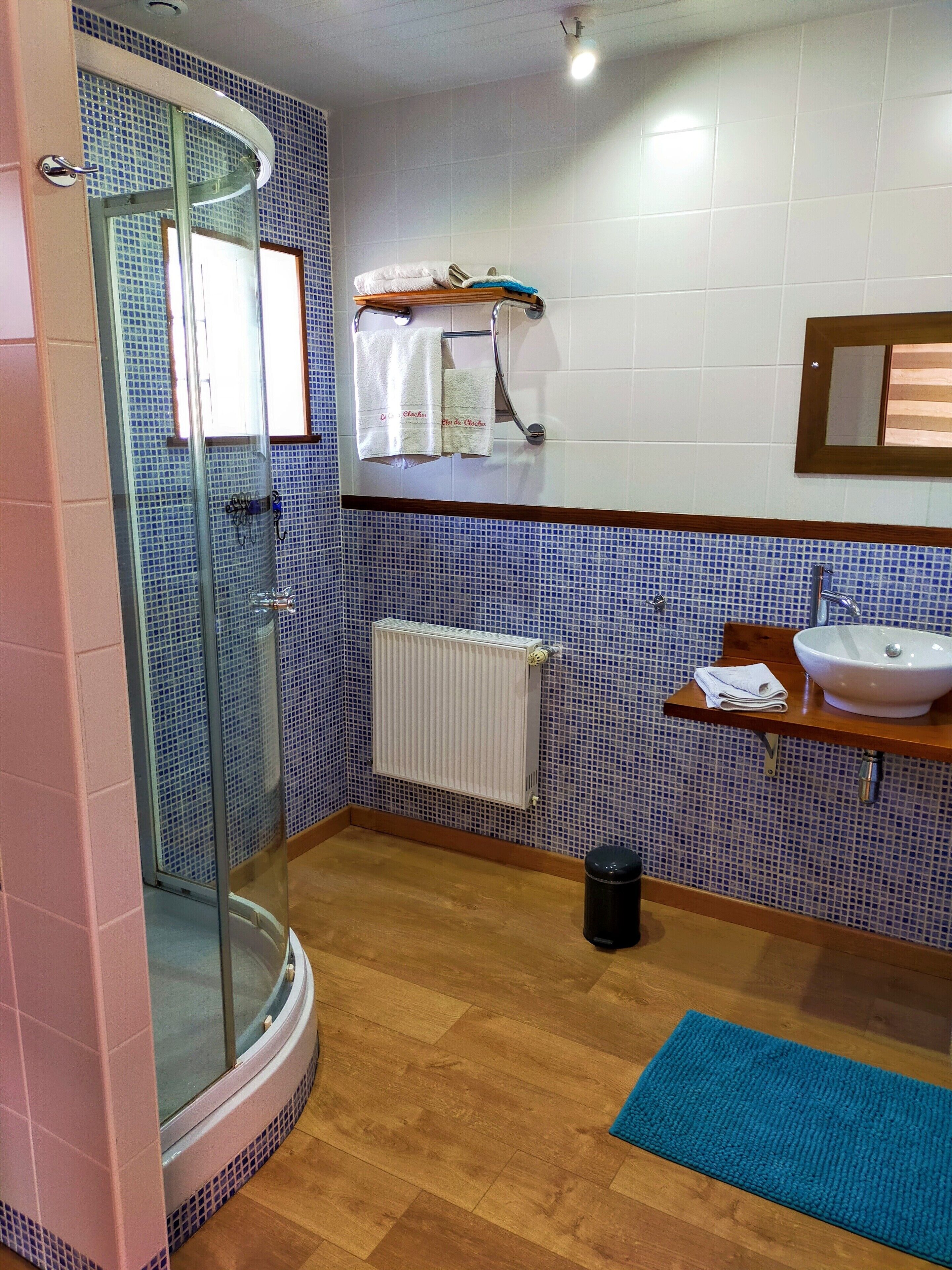 Un baño con cabina de ducha de vidrio, un lavamanos blanco, un perchero con toallas, un radiador y un felpudo azul en el suelo.
