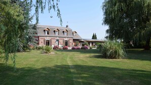 A large brick house with a sloped roof, surrounded by a well-maintained lawn and flowering plants. - Le Clos du Clocher (Gueudecourt)
