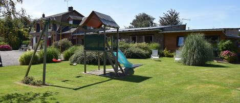 Un parque infantil con tobogán y columpios, rodeado de un césped cuidado y un edificio de ladrillo con techo inclinado.