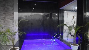 A modern indoor water feature with a glass enclosure, illuminated by blue lighting, and surrounded by potted plants. - Hotel Royal Country Club (Pindra)