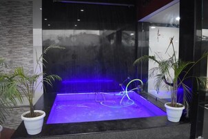 Una fuente de agua moderna en interior, con recinto de vidrio, iluminada con luces azules y rodeada de plantas en macetas.