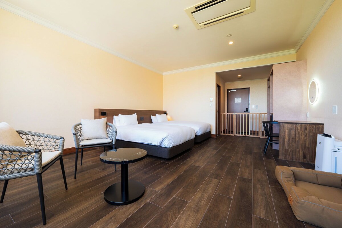 Quarto de hotel com cama, duas cadeiras, uma mesinha e uma escrivaninha com computador.