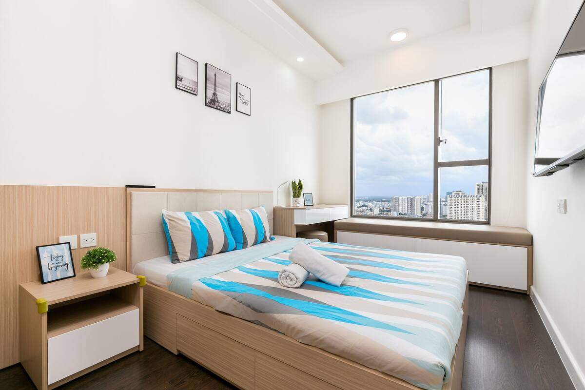 A modern bedroom with a large bed, a nightstand, a small table with a plant, and a window with a city view.