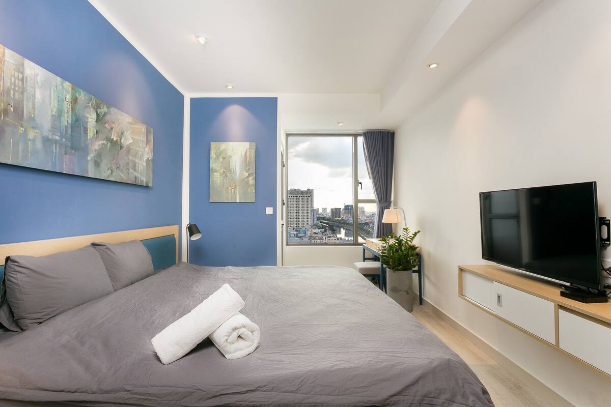 A modern hotel room with a large bed, a flat-screen TV, a desk with a lamp, and a city view through the window.
