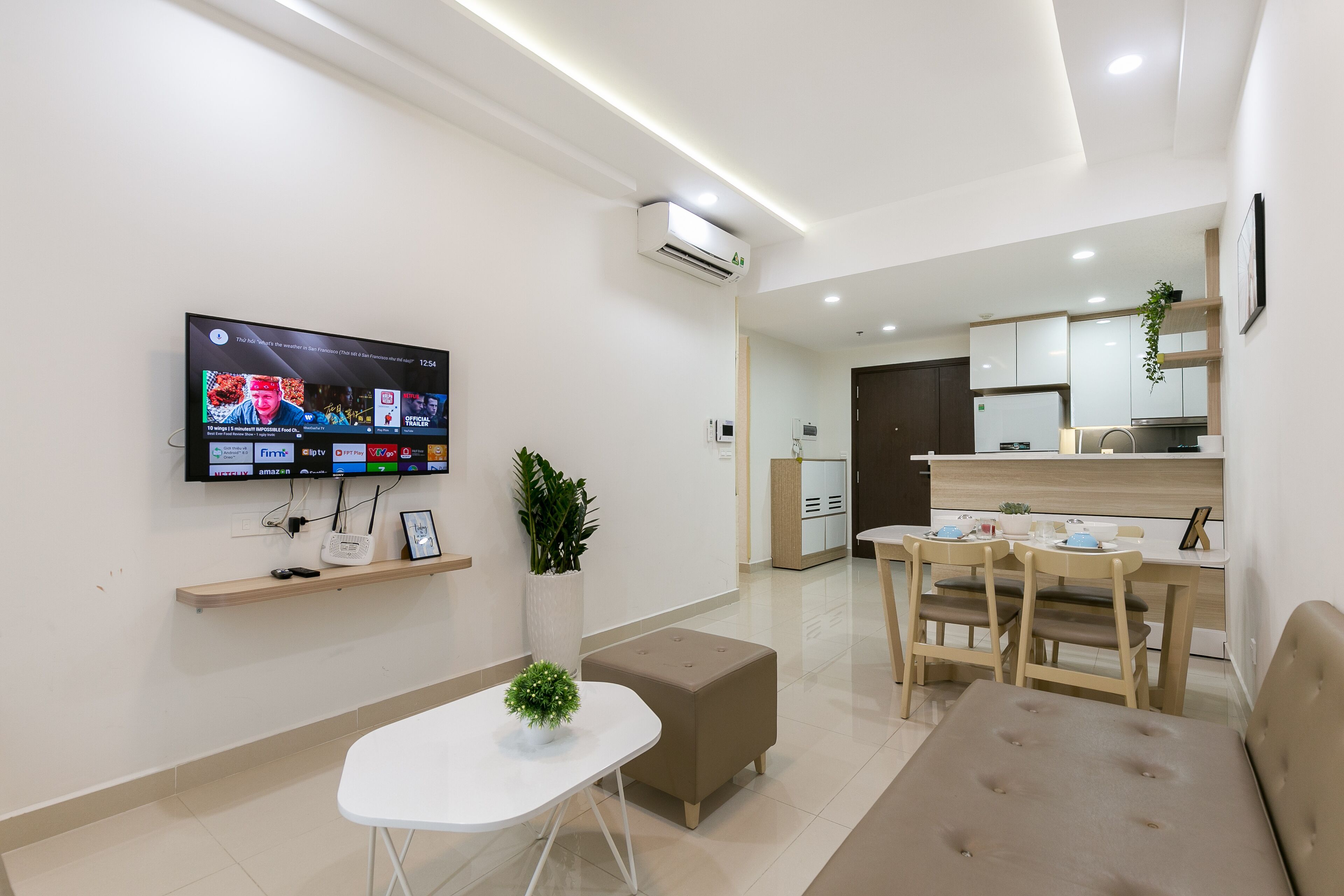 Uma sala de estar moderna com televisão de tela plana, uma mesa pequena, um sofá e um espaço de jantar com cadeiras, além de uma cozinha ao fundo.