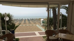A balcony with wicker furniture, a view of a beach with rows of sun loungers, and a small structure on the sand. - Hotel Ariston (Caorle)