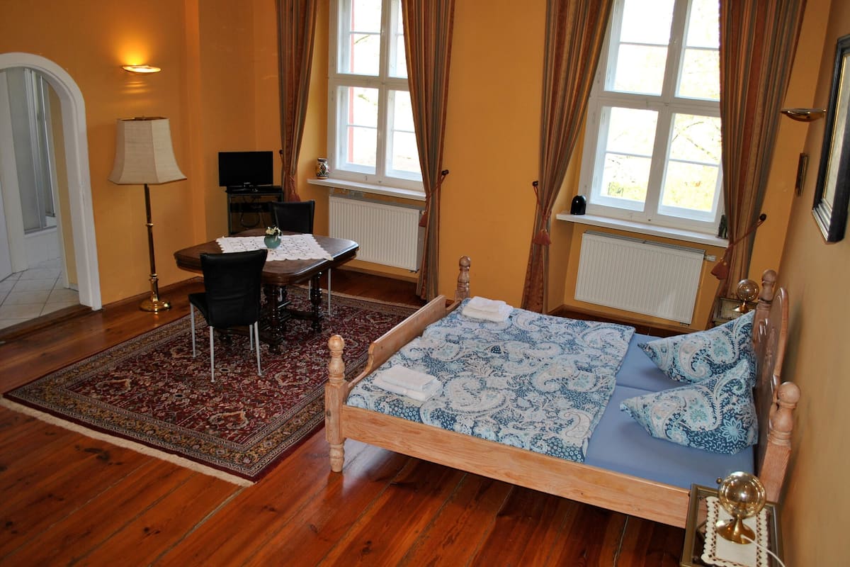 A bedroom with a wooden bed, a desk, a chair, a lamp, and a view of the outdoors through large windows.