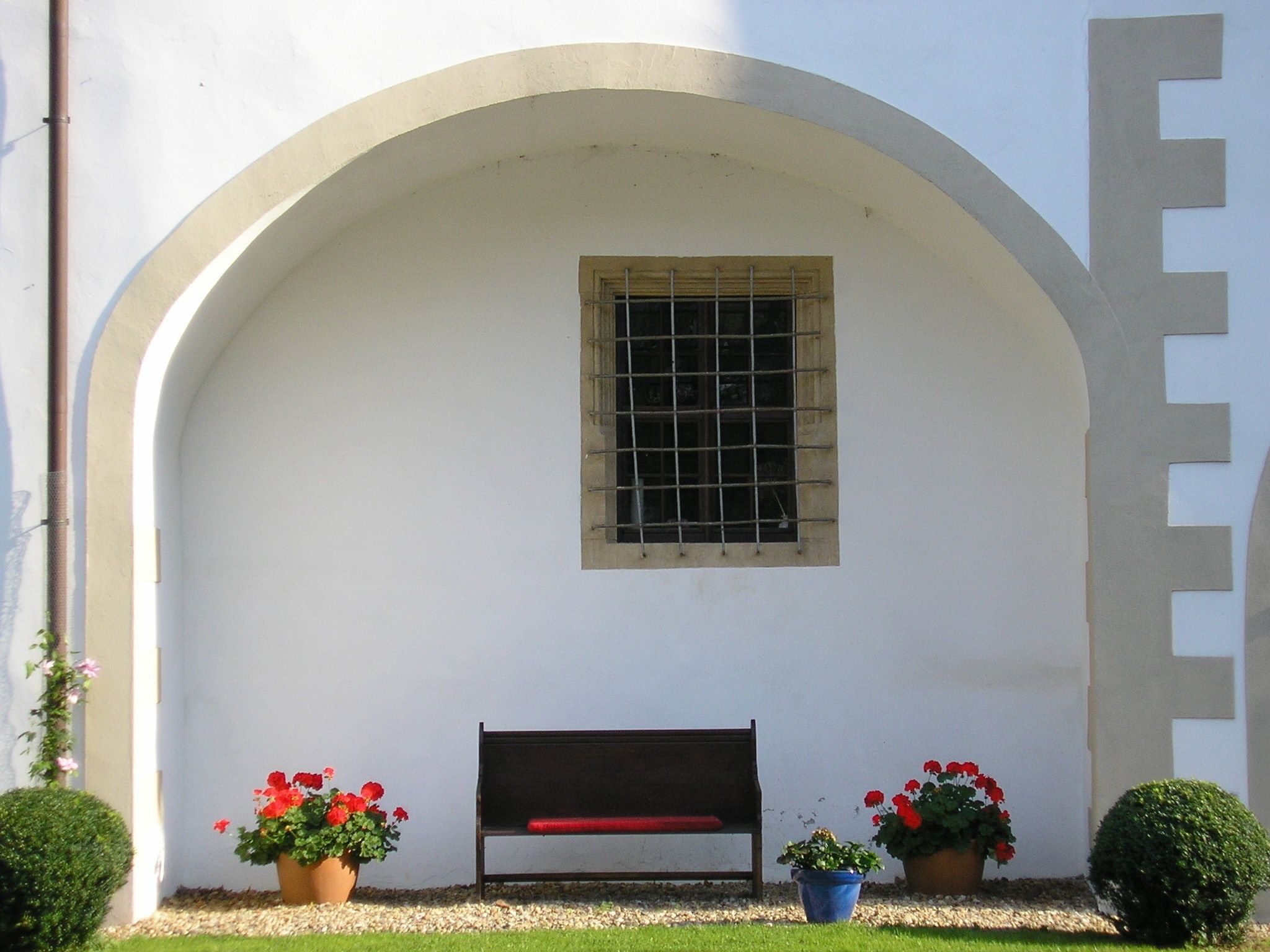 A white building with a large arched doorway, a window with metal bars, and a bench in front. There are potted plants with red flowers on either side of the bench.