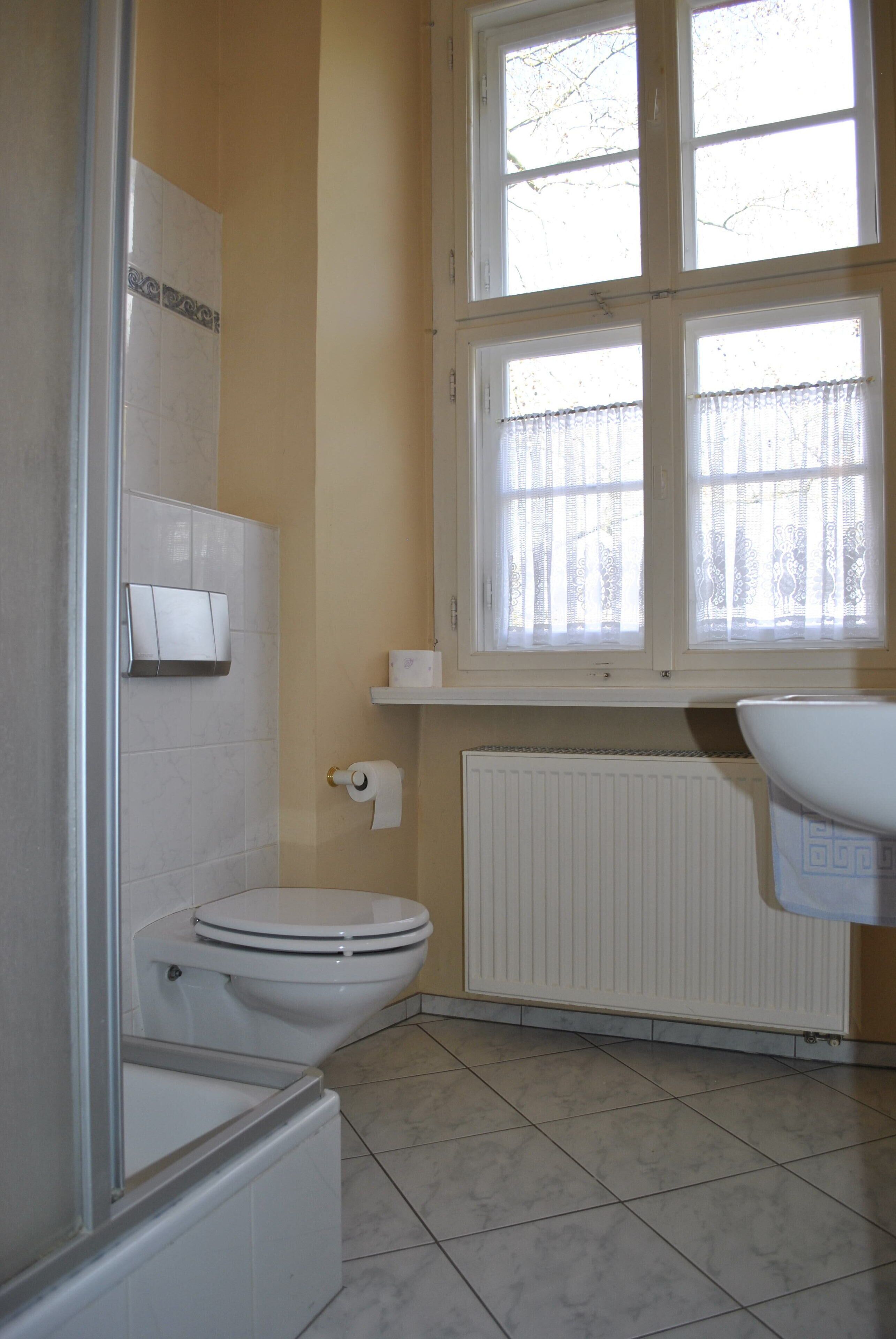 A bathroom with a toilet, a shower, a radiator, and a window with curtains.