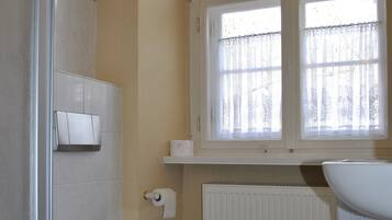 A bathroom with a toilet, a shower, a radiator, and a window with curtains.