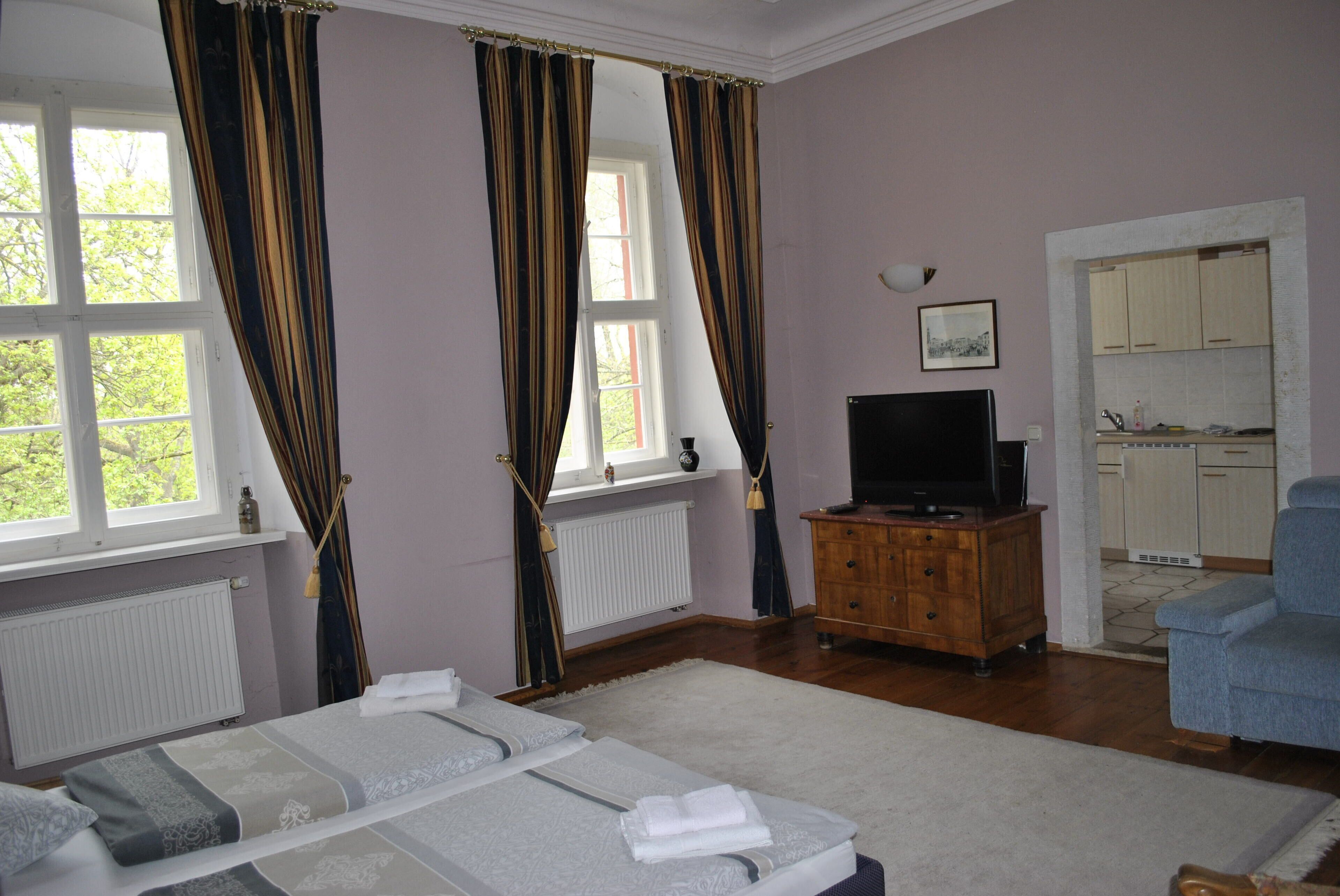 Quarto com cama, televisão, cómoda de madeira, cadeira e vista para a cozinha.