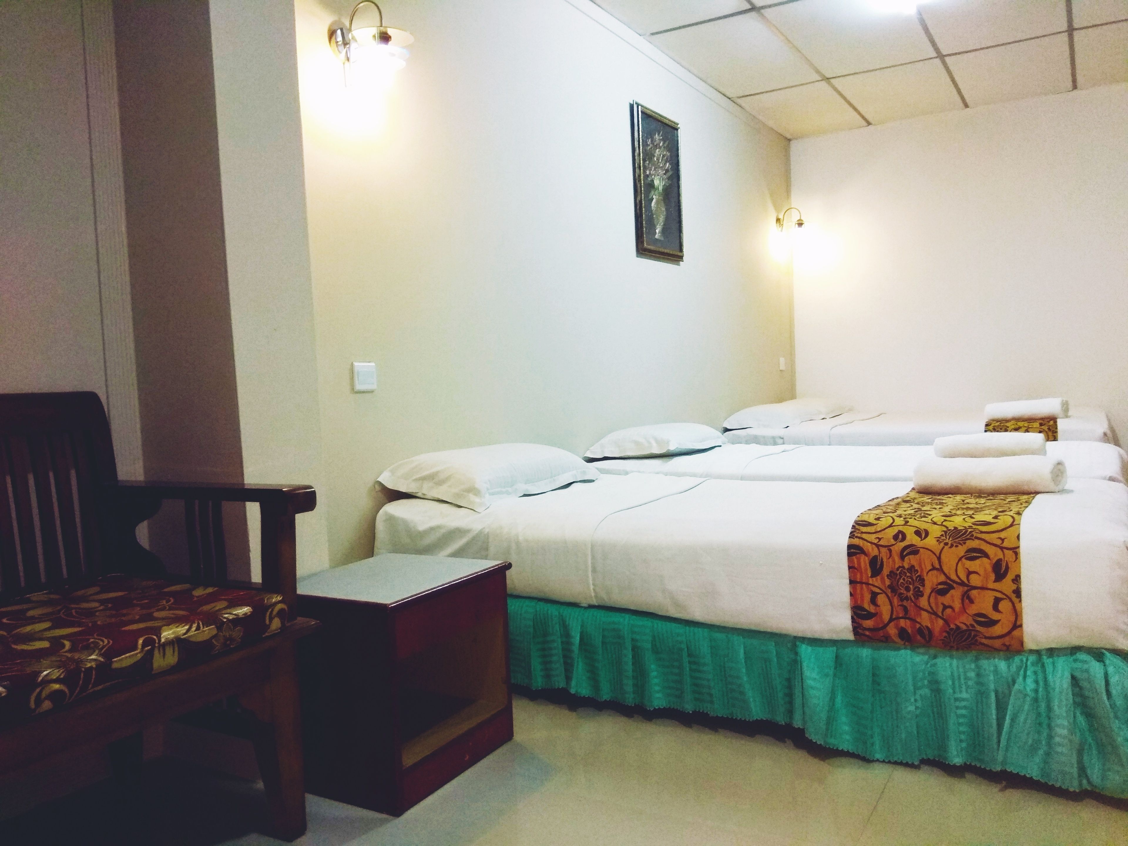 A hotel room with two beds, a wooden chair, a small table, and a green skirted bed.
