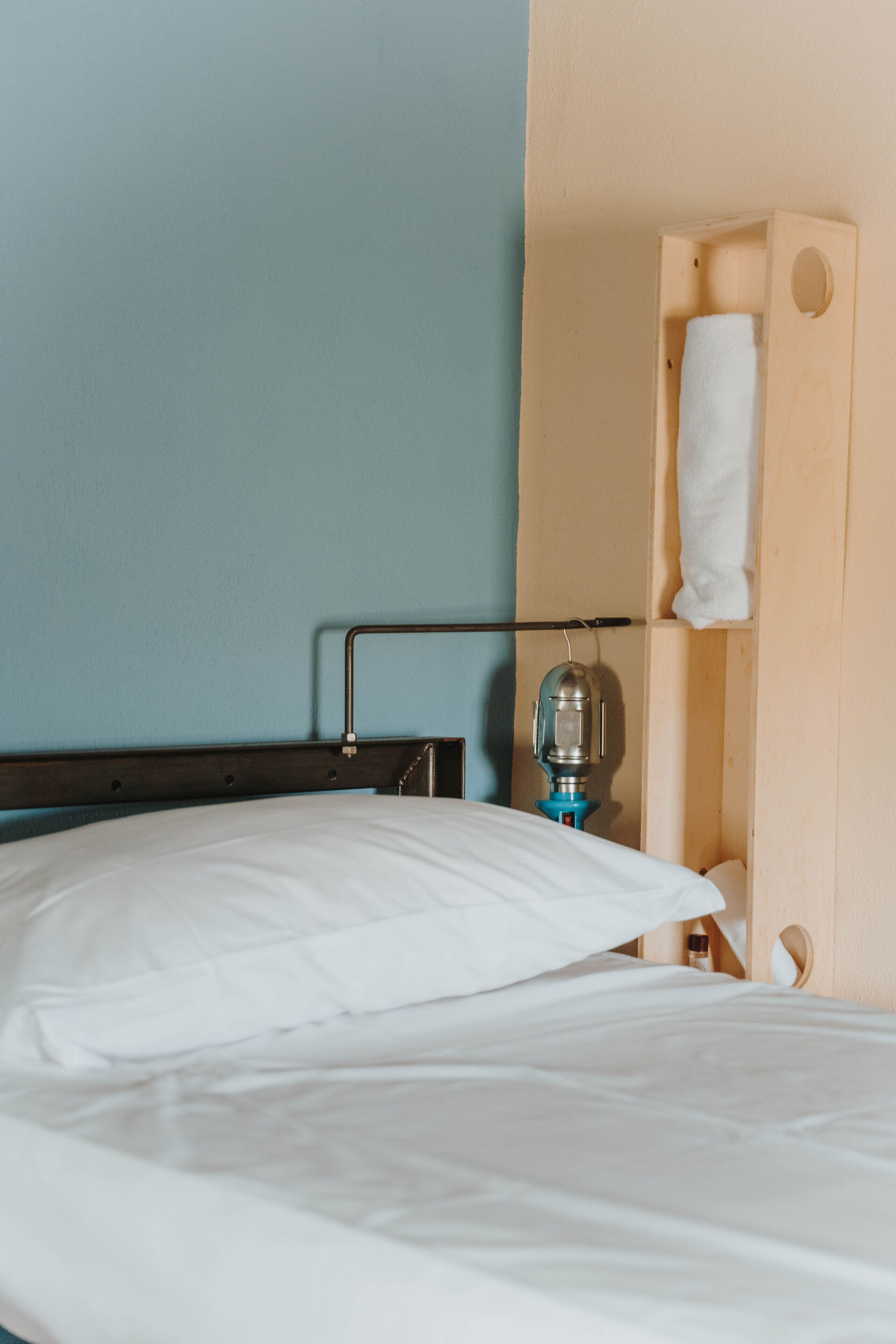 Un lit avec des draps blancs, une tête de lit en bois, une lampe fixée au mur et une serviette suspendue à une étagère en bois.