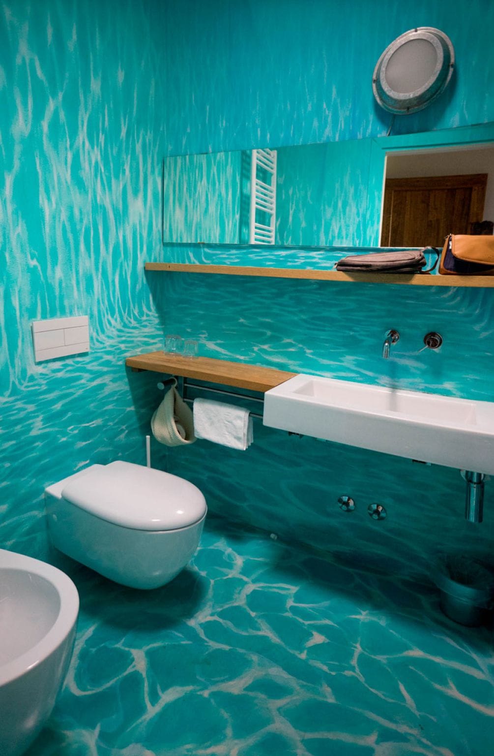 A bathroom with a turquoise wall and floor, a wooden vanity, a white sink, and a toilet.