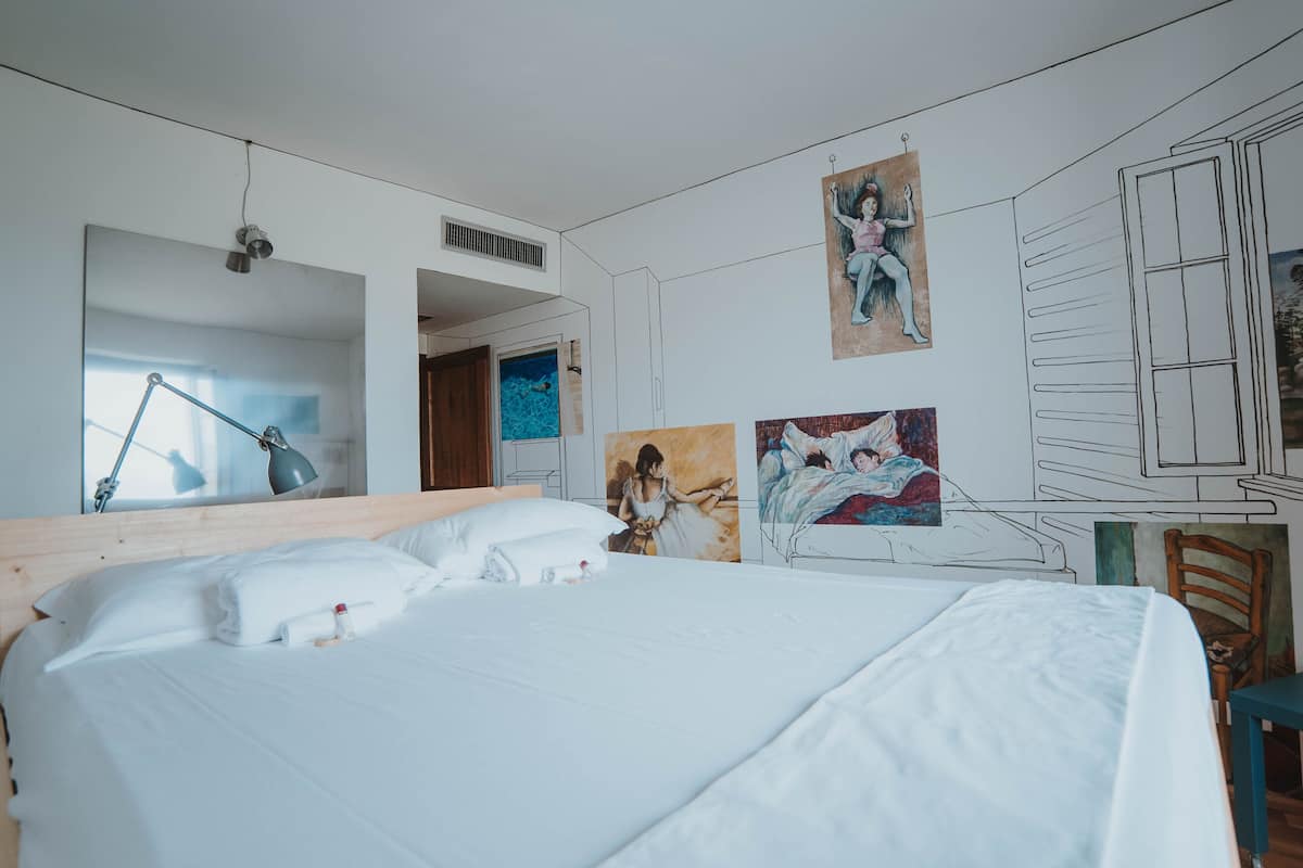A hotel room with a large bed, a desk, and a chair. There are paintings on the walls and a window with a view of the sea.