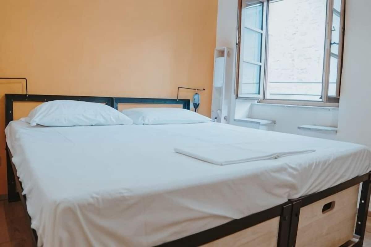 A single bed with white bedding, a wooden headboard, a bedside table, and a window with a view of the outside.