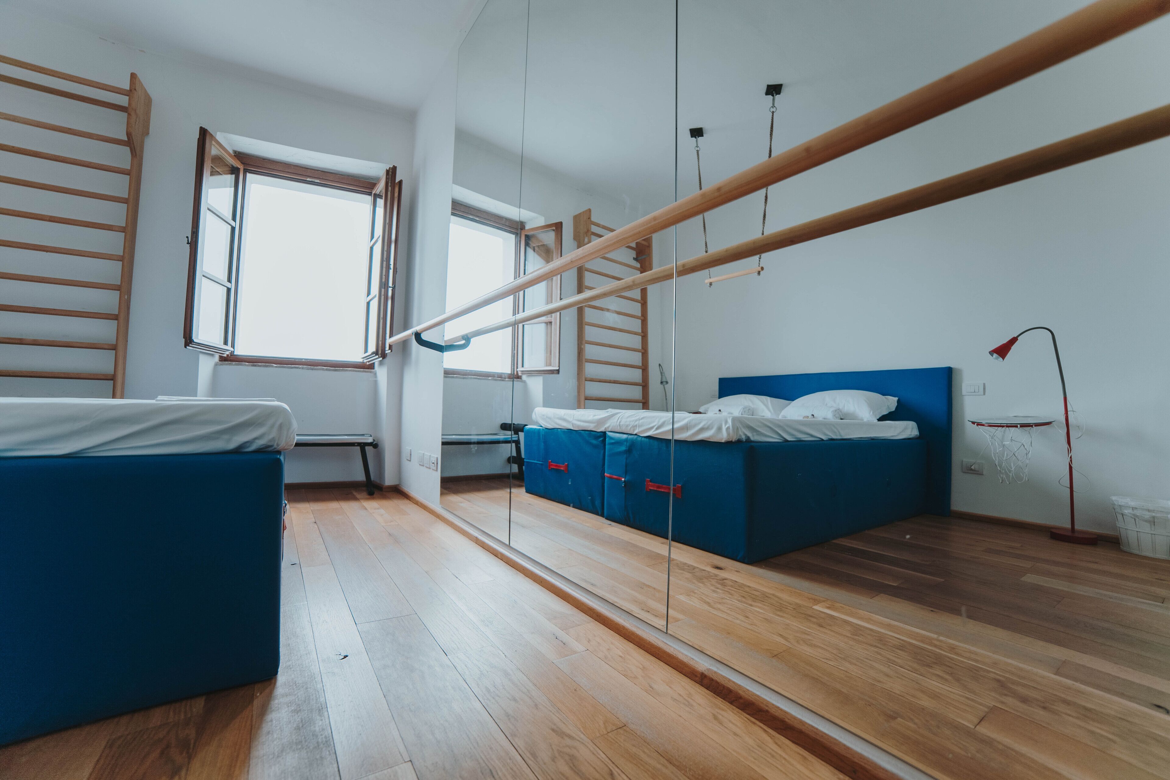 Una habitación con piso de madera, una cama con cabecera azul, un escritorio pequeño, una escalera y una ventana.