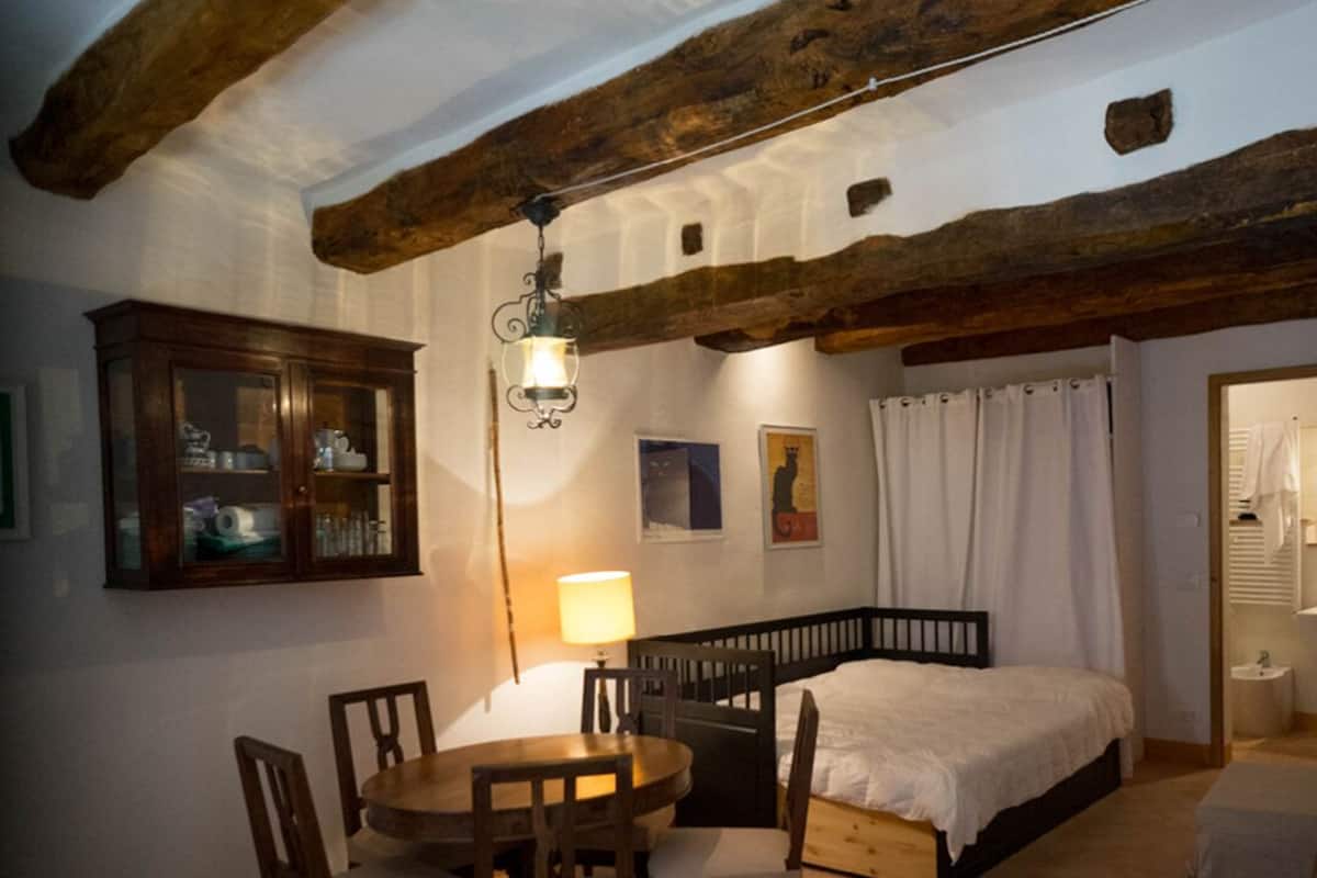 A cozy bedroom with a wooden headboard bed, a dining table with chairs, a cabinet with glass doors, and a hanging light fixture.