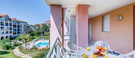 A balcony with a white table set for breakfast, overlooking a swimming pool and lush greenery.
