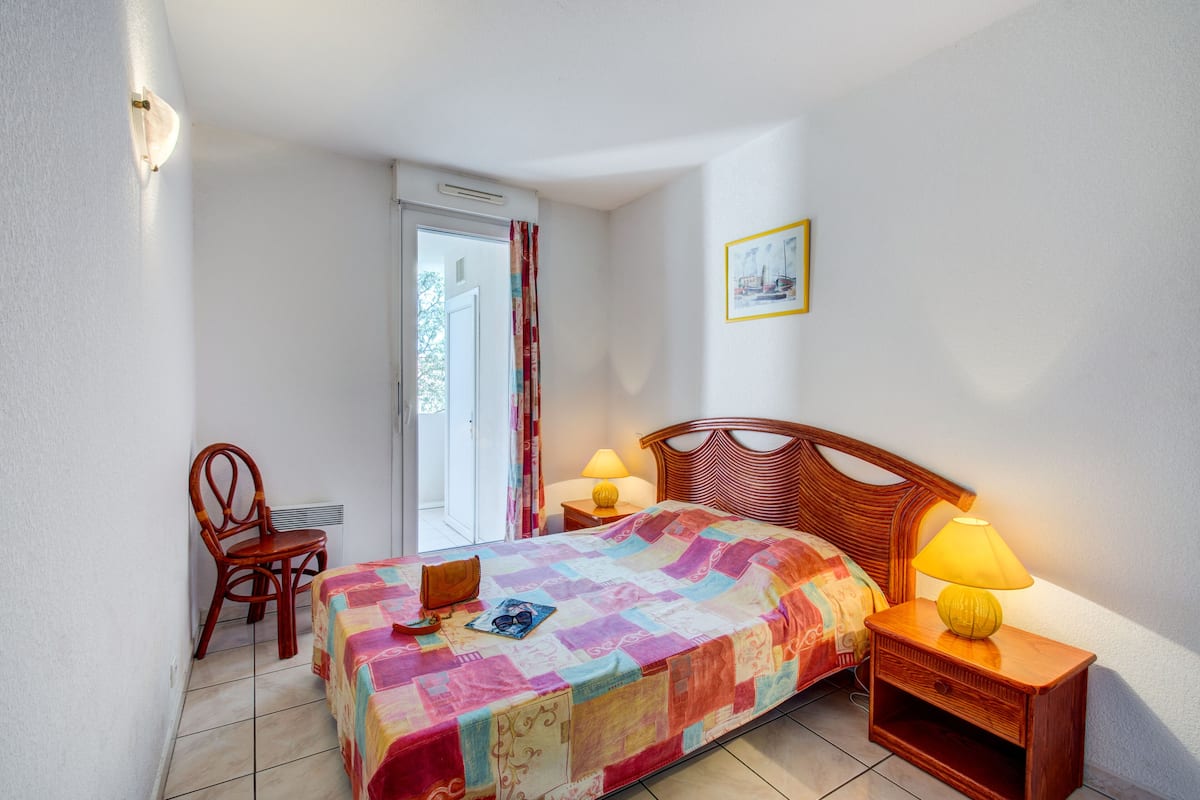 Un dormitorio con cama, una mesita de noche de madera con lámpara, una silla y una puerta que da a un balcón.