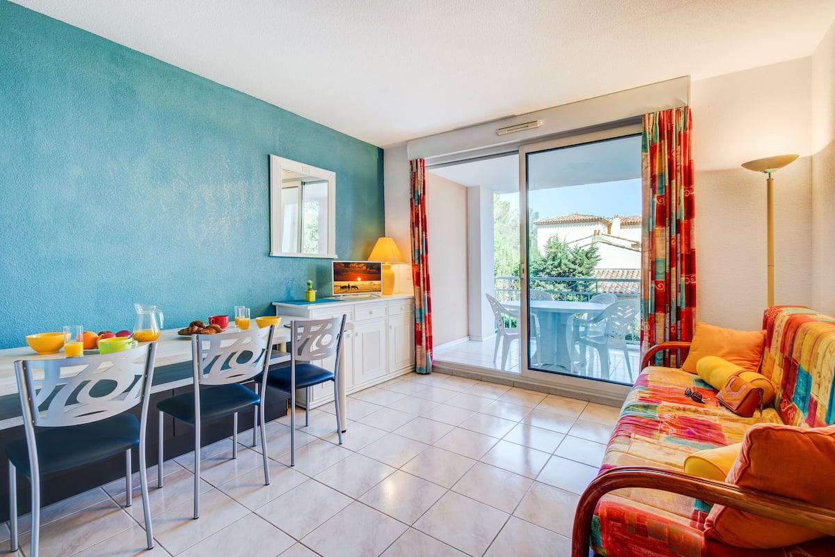 Un salón luminoso con una mesa de comedor preparada para el desayuno, un sofá con una manta colorida y vistas a un patio exterior a través de puertas correderas de cristal.
