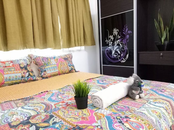 Un dormitorio con una colcha colorida, una toalla enrollada, un peluche, una pequeña planta y un adorno de pared.
