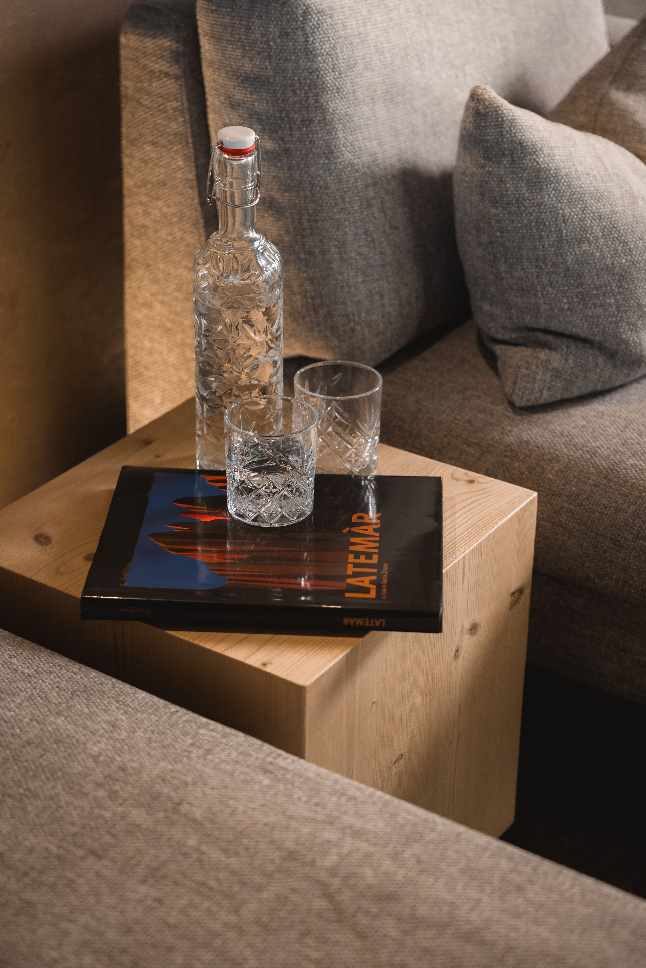 Una mesita de madera con una botella de vidrio, dos vasos y una revista sobre un cojín.