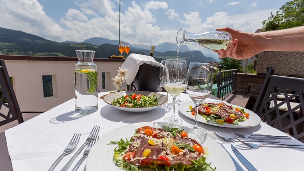 Une table dressée pour un repas, avec une vue sur la montagne, une bouteille d’eau et deux verres de vin blanc.