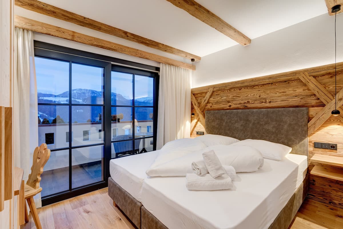 A modern hotel room with a large bed, wooden ceiling beams, a view of mountains, and a sliding glass door to the balcony.