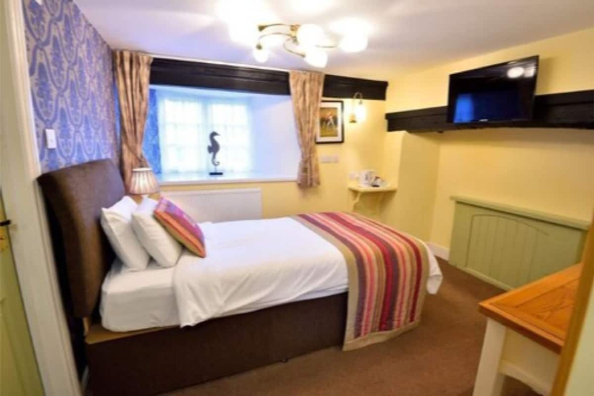 Une chambre à coucher avec un lit, une fenêtre avec des rideaux, un téléviseur fixé au mur, une table de chevet et un radiateur.