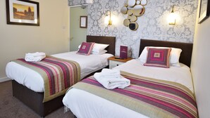 A hotel room with two beds, each with a striped blanket and a pillow. There is a nightstand with a lamp and a mirror. The room has a patterned wallpaper and a framed picture on the wall. - The Crown Hotel (Hailsham)