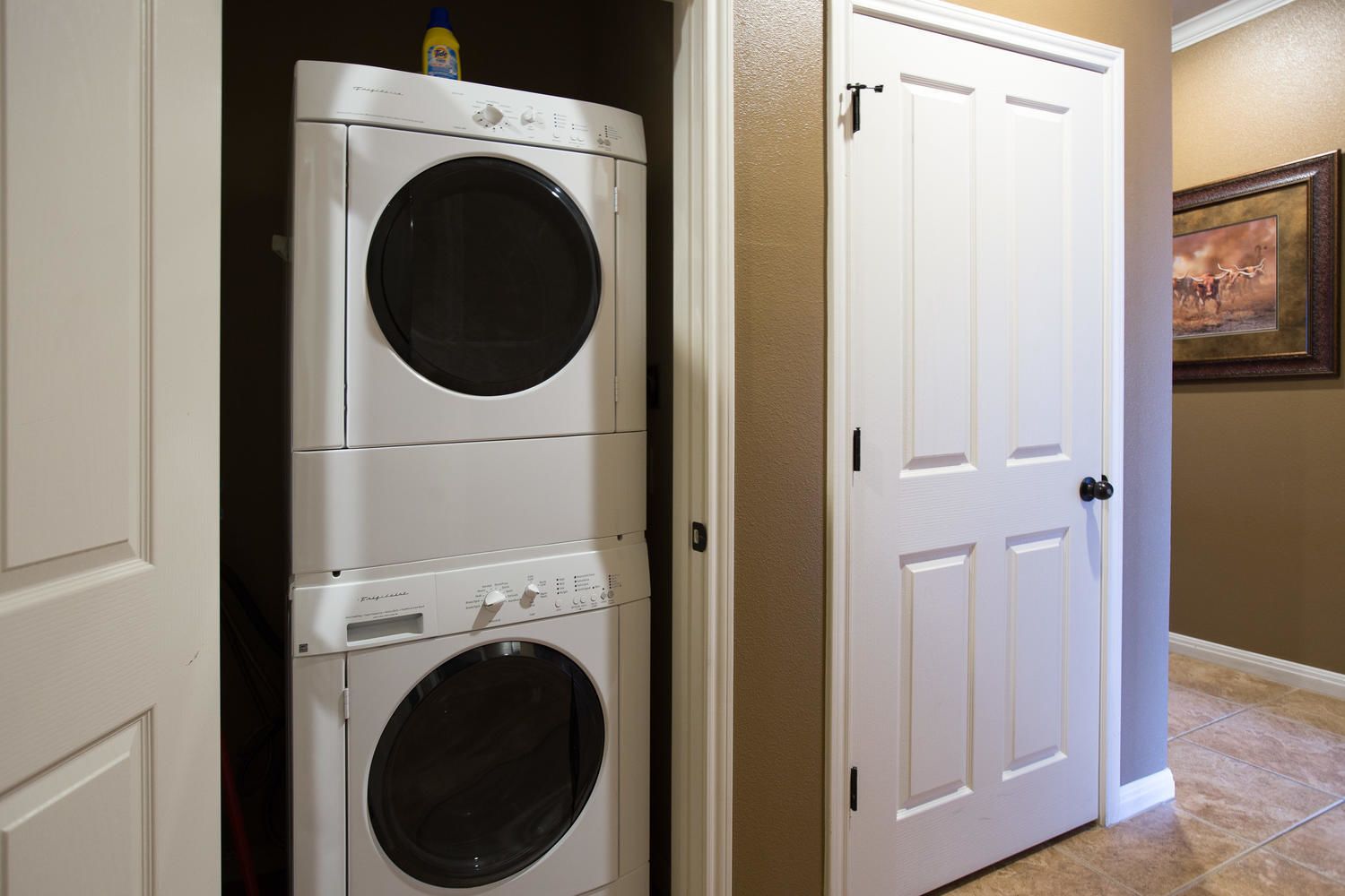 Une buanderie équipée de deux machines à laver et deux sèche-linge à chargement frontal, une porte blanche et un tableau encadré au mur.