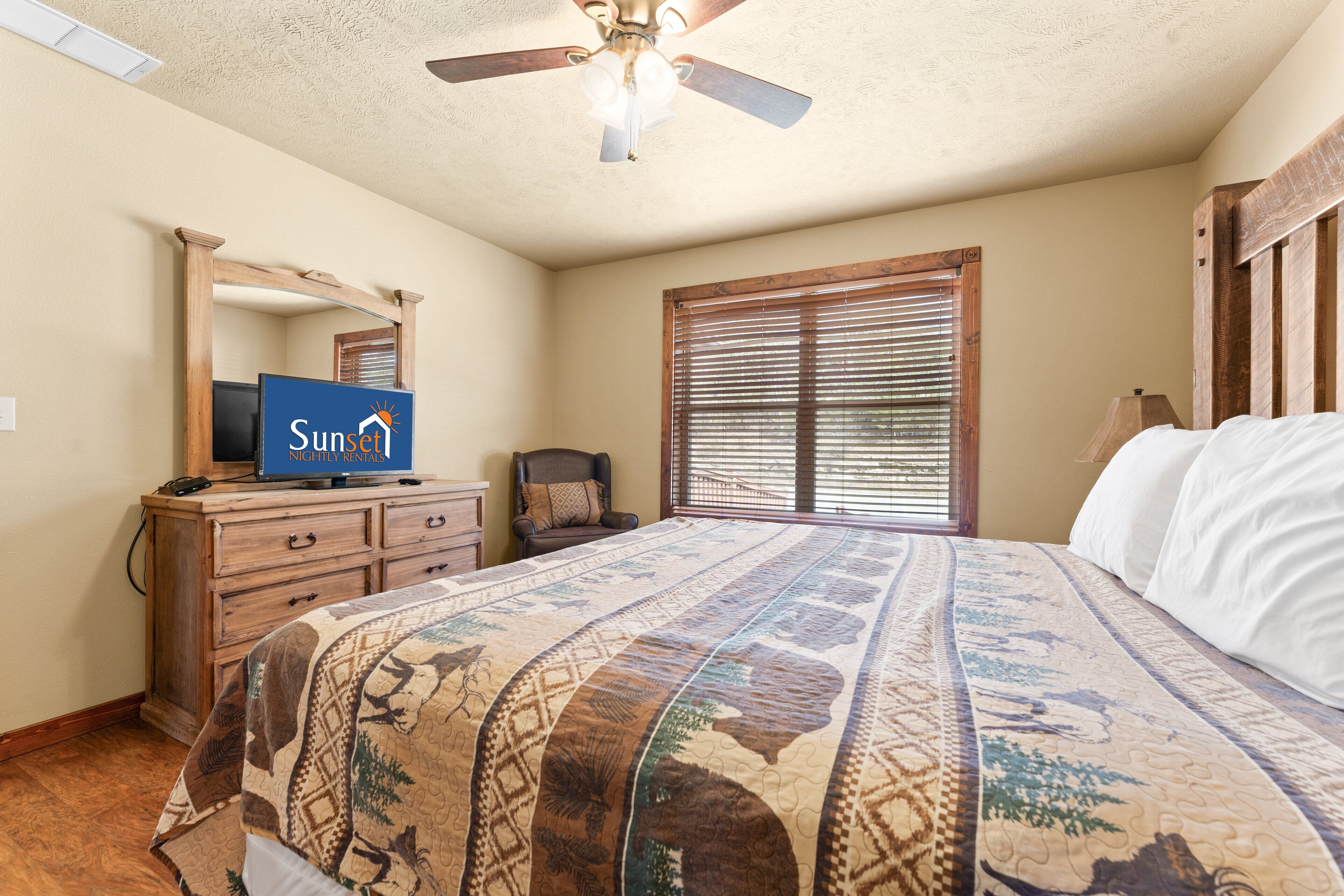 A bedroom with a bed, a ceiling fan, a wooden dresser, a television, and a window with blinds.