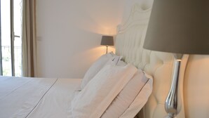 Ein ordentlich bezogenes Bett mit weißen Leinen und Kissen, eine Nachttischlampe und ein Blick ins Freie durch ein Fenster mit Vorhängen.