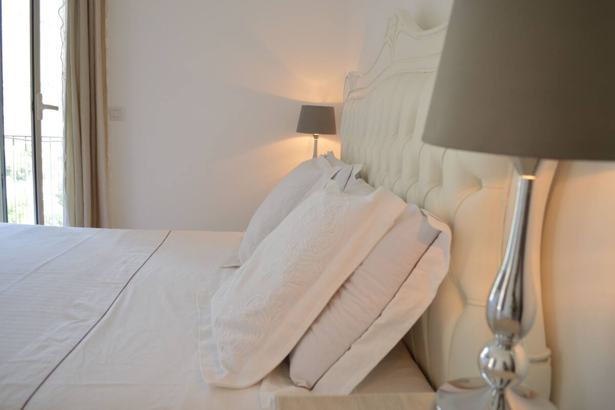 A neatly made bed with white linens and pillows, a bedside lamp, and a view of the outdoors through a window with curtains.