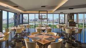 A modern dining area with tables and chairs, large windows with a city view, and hanging light fixtures.