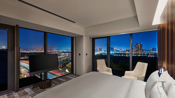 A hotel room with a large bed, two white armchairs, a flat-screen TV, and large windows offering a city view.