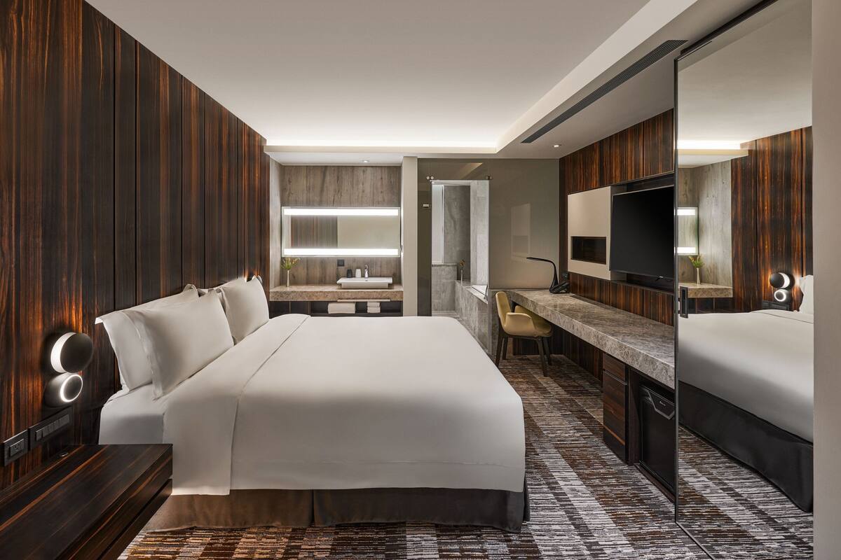A modern hotel room with a large bed, a desk, a TV, and a bathroom with a mirror and sink.