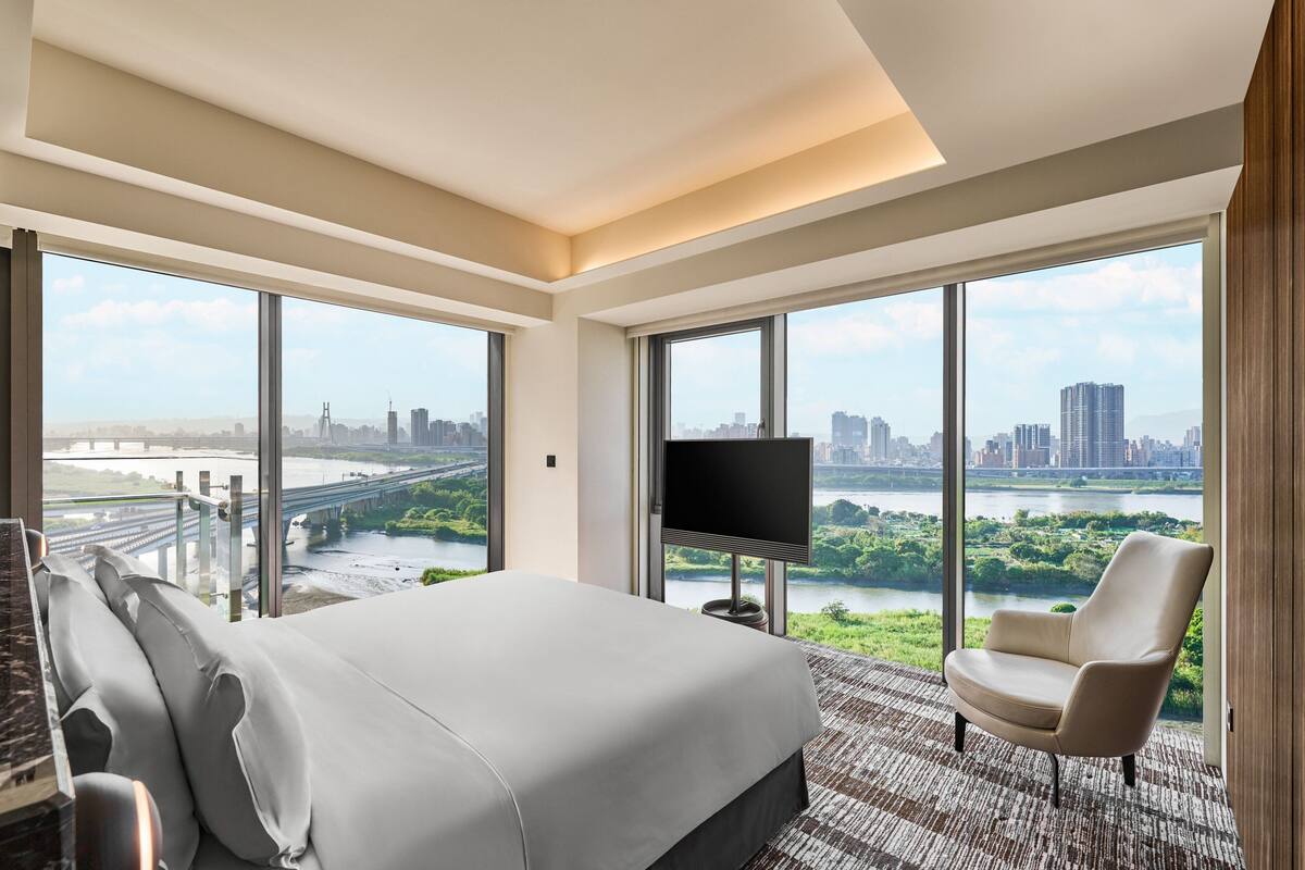 A spacious bedroom with a large bed, a TV, and a chair, featuring a cityscape and river view through large windows.
